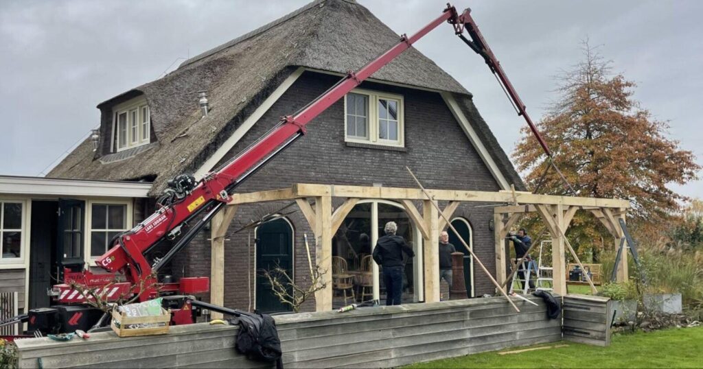 Mini hijskraan plaatst een houtconstructie bij een woning met rieten dak
