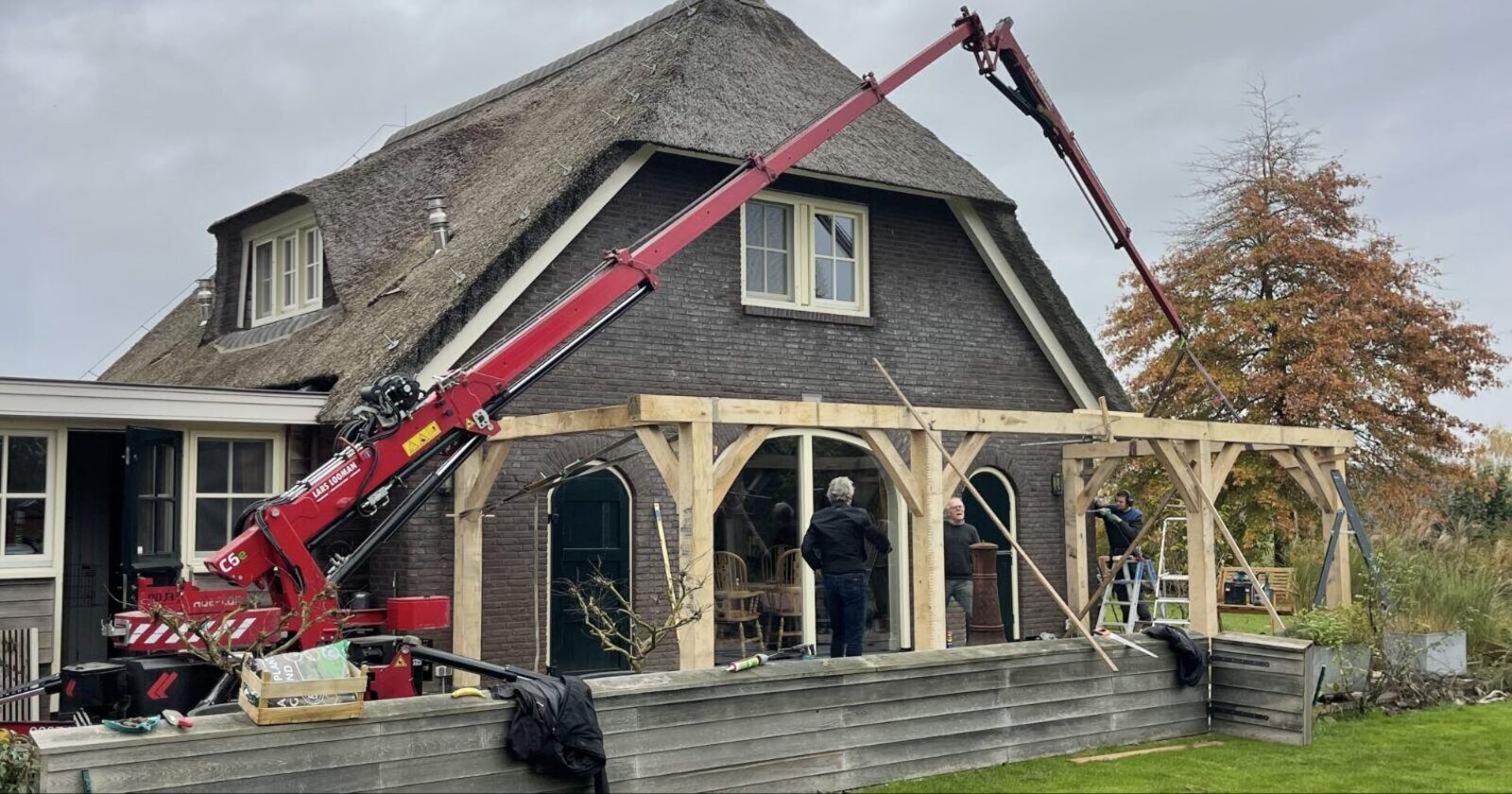 Mini hijskraan plaatst een houtconstructie bij een woning met rieten dak